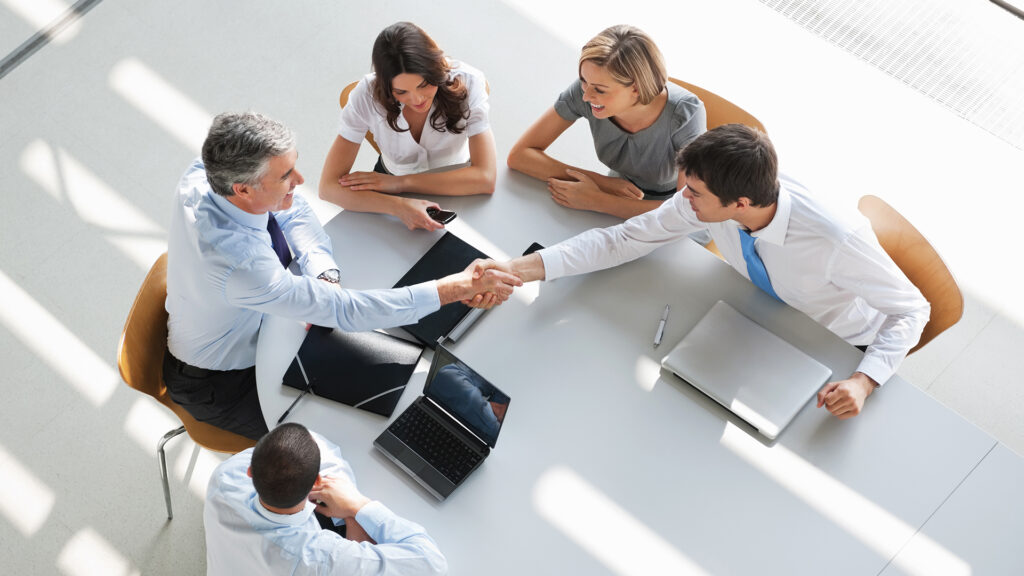 Business meeting with handshake between two executives, two colleagues observing.