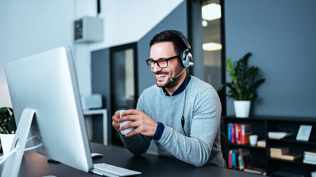 A man with a headset, smiling while holding a coffee cup and sitting at a desk with a computer monitor