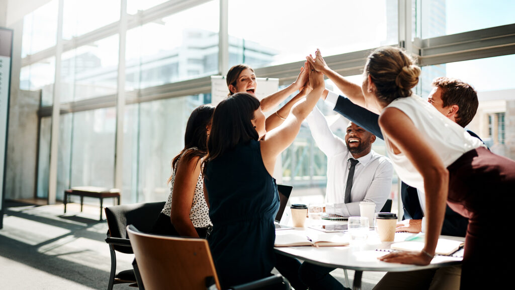 Business team members high-fiving around a table in a brightly lit office space.