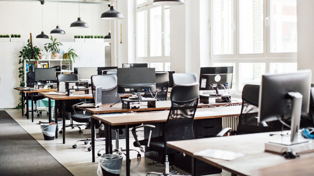 An empty office space with multiple workstations, chairs, computers, and large windows, indicating a modern office environment.