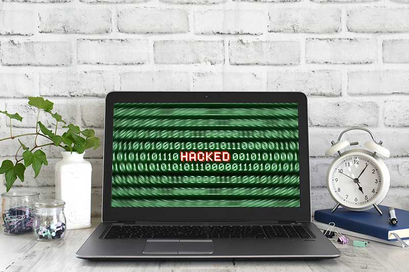 Close-up view of a laptop screen displaying a “HACKED” warning message in red text amidst binary code, surrounded by a home office setup with plants, an alarm clock, and stationery.