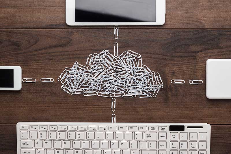 Paperclips arranged in the shape of a cloud connect to a keyboard, smartphone, and tablet on a wooden surface.