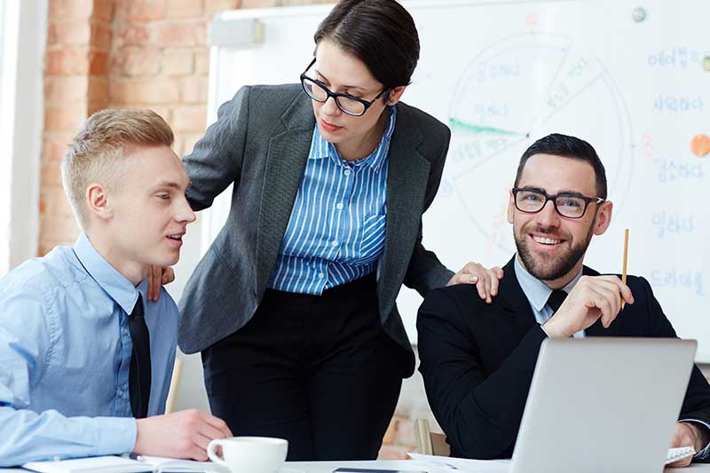 A businesswoman stands over two male colleagues, engaged in a meeting with a laptop and coffee cups on the table, suggesting collaboration.