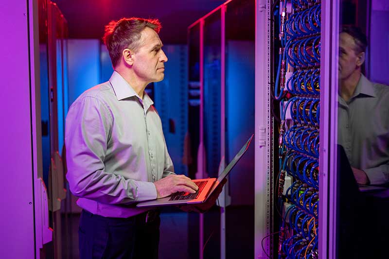 Man in a server room working on a laptop, surrounded by colorful lighting and server racks.