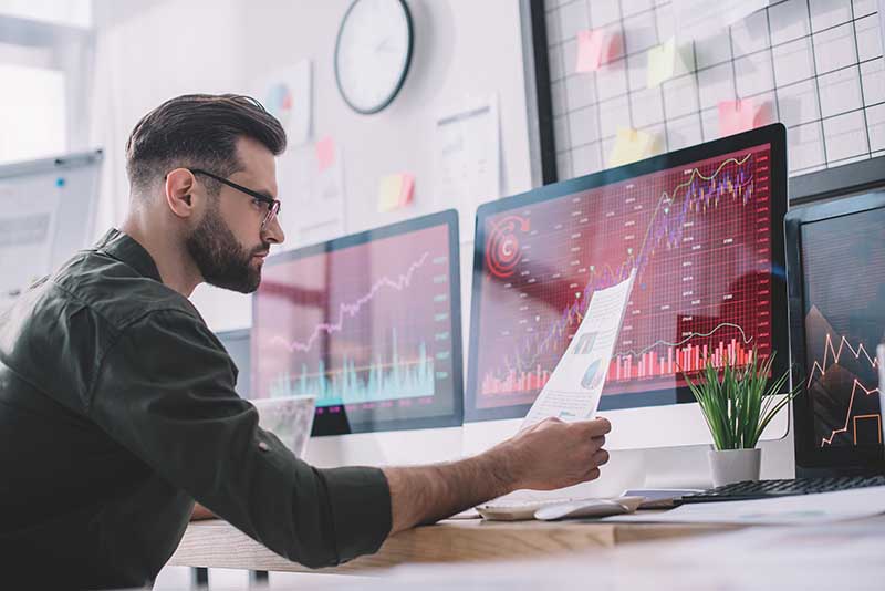 A man with a beard and glasses reviews financial charts on multiple computer screens. 5G