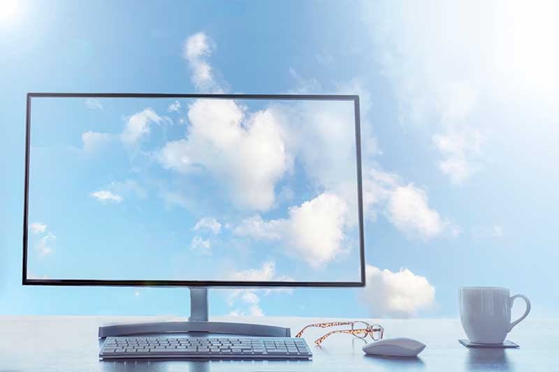 Computer monitor showing a sky with clouds, symbolizing cloud computing. The desk includes a keyboard, mouse, glasses, and a coffee cup.