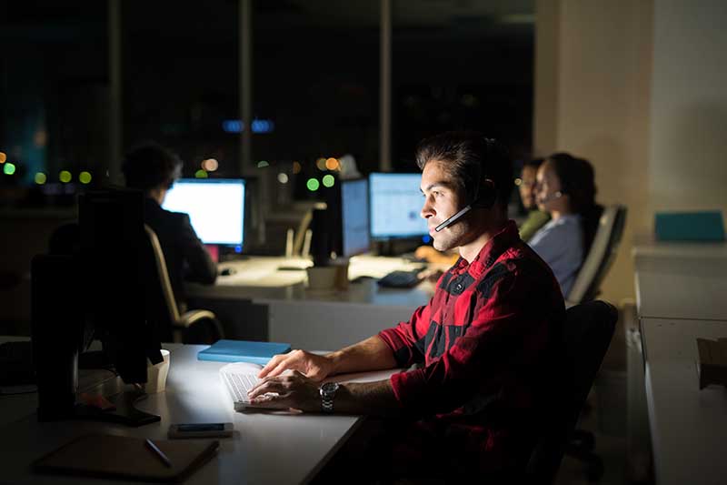 A man wearing a headset and working at a computer in a dimly lit office, with two other people working in the background.