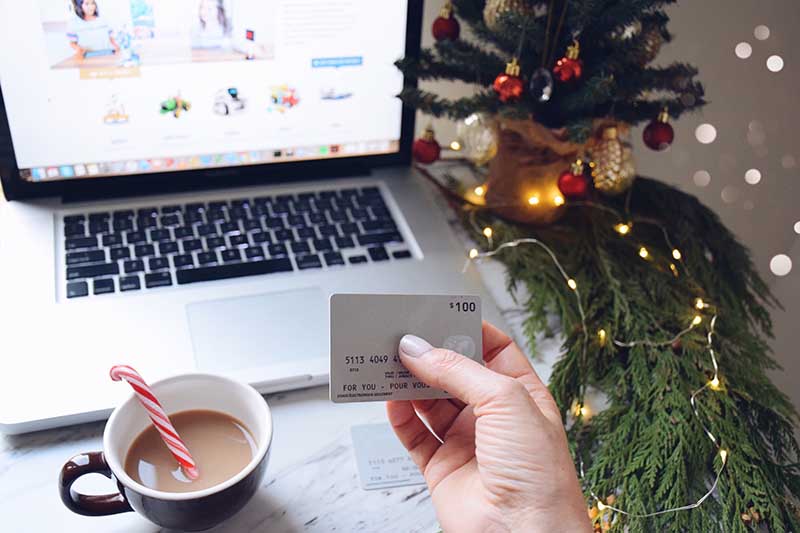 A hand holding a credit card in front of a laptop with a cup of coffee and Christmas decorations nearby.