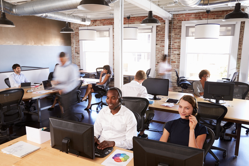 Employees working at desks with computers and headsets in a modern open-plan office with exposed brick walls and large windows.