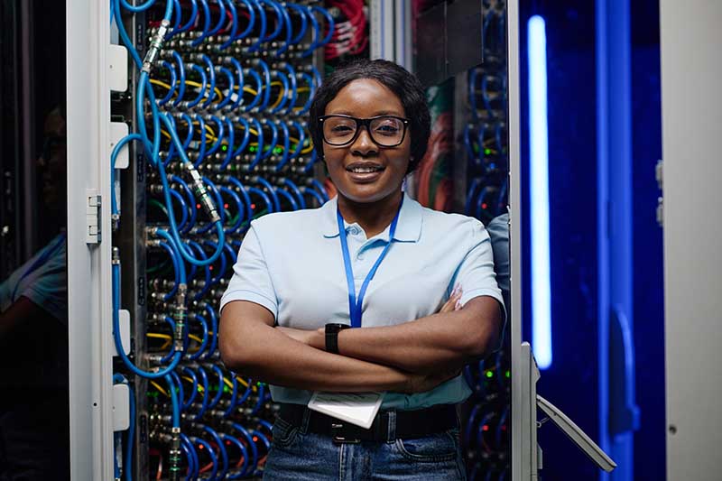 Woman standing confidently in front of a server rack, arms crossed, wearing glasses and a light blue shirt.