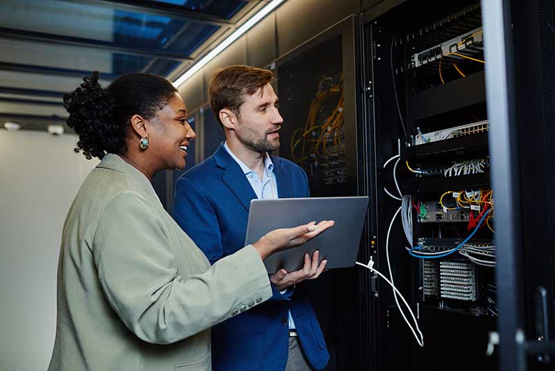 Two people in a data center discussing server management while looking at a laptop