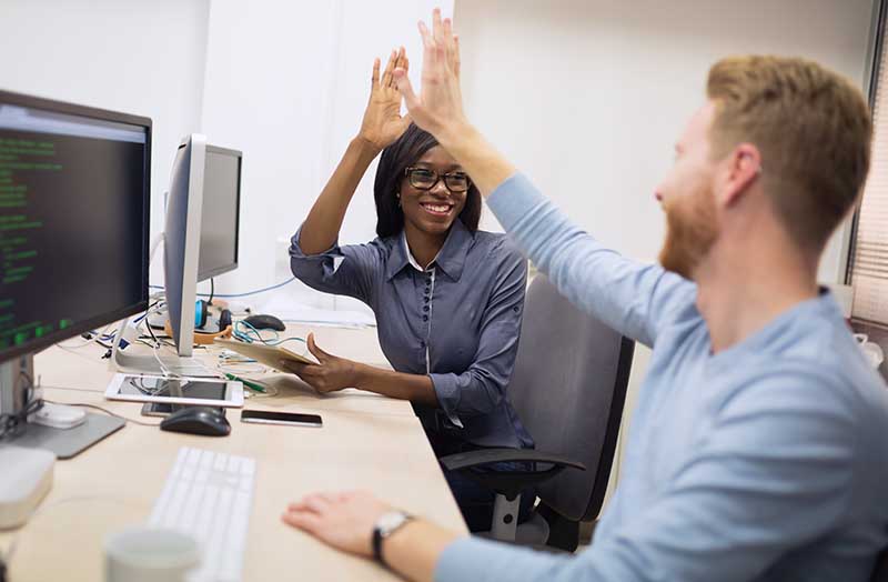 Two colleagues high-fiving each other at a desk with computer monitors and other office equipment.