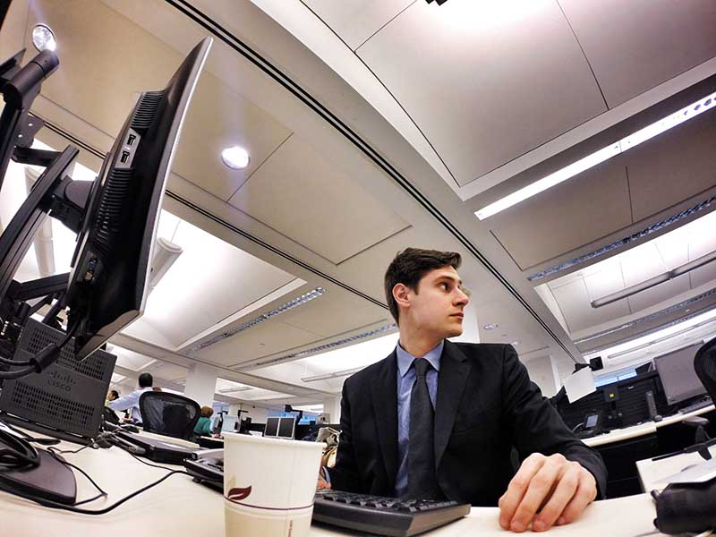 A young professional in a business suit sits at a desk, looking off to the side while working in a modern office with multiple computer monitors.