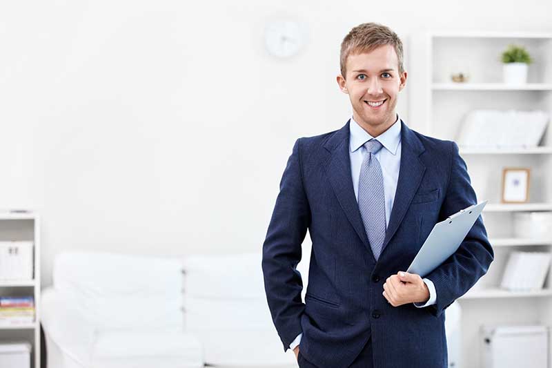 Man in a business suit holding a clipboard, standing in a modern office.