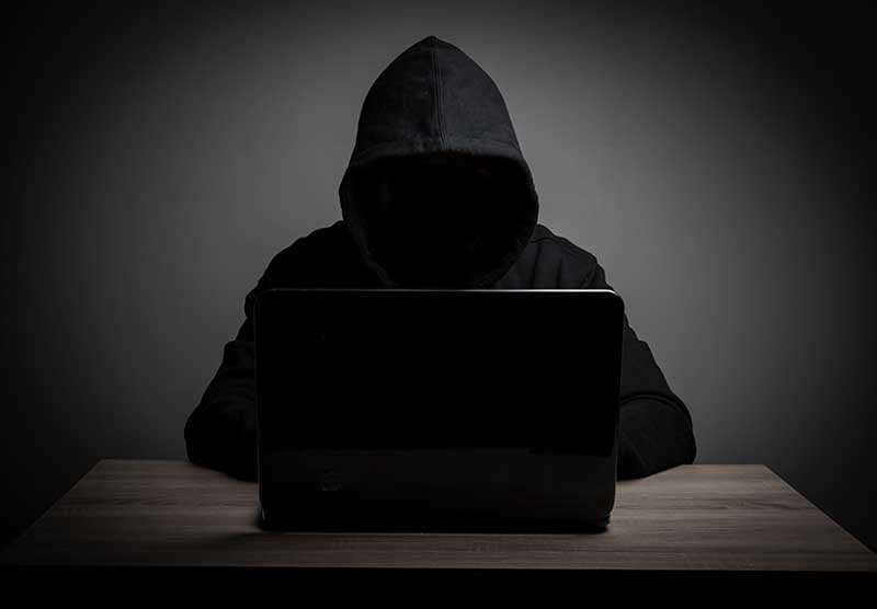 A person in a dark hoodie sits in front of a laptop in a dimly lit room, creating a mysterious and ominous atmosphere.