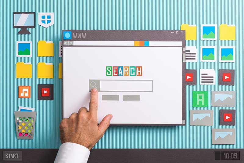 A hand pointing at a search bar on a paper cutout illustration of a web browser with various digital icons in the background.