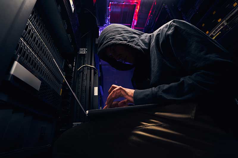 A hooded figure working on a laptop inside a server room.